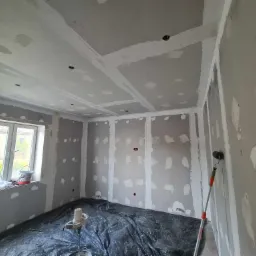 Pokój w trakcie prac wykończeniowych: ściany i sufit z płyt kartonowo-gipsowych, z widocznymi łączeniami i miejscami po szpachlowaniu, zabezpieczona podłoga folią, widoczny wałek malarski oparty...