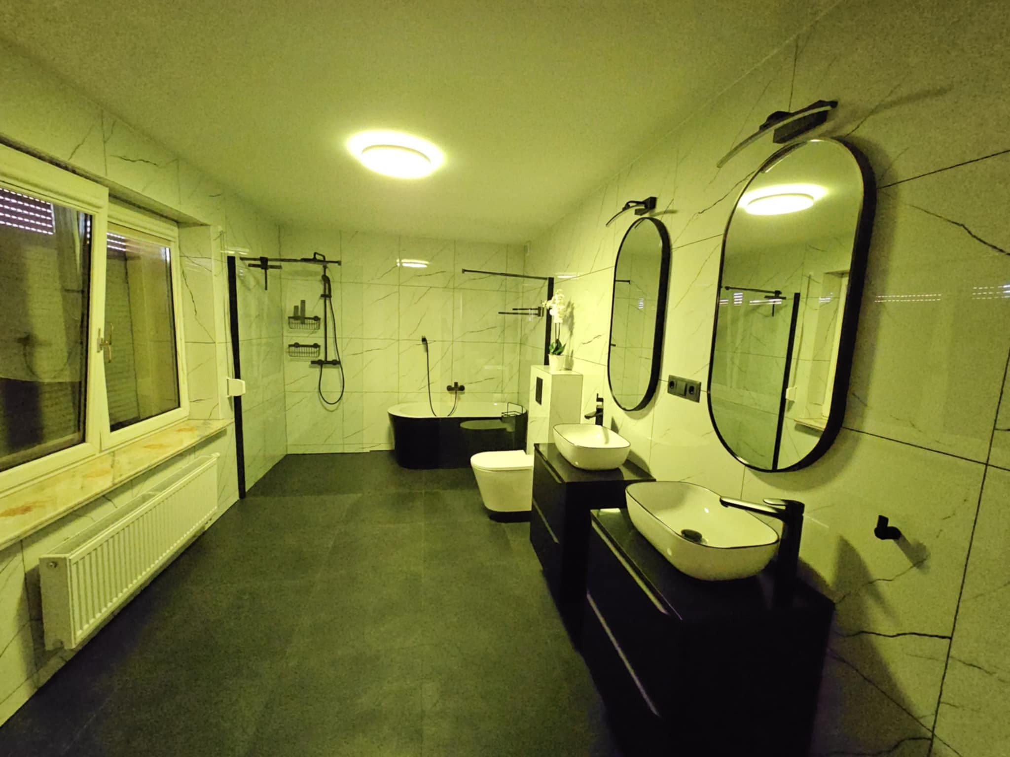 Wykończona łazienka z czarną wanną wolnostojącą, białymi umywalkami nablatowymi na czarnych szafkach i marmurowymi płytkami na ścianach, oświetlona światłem LED.