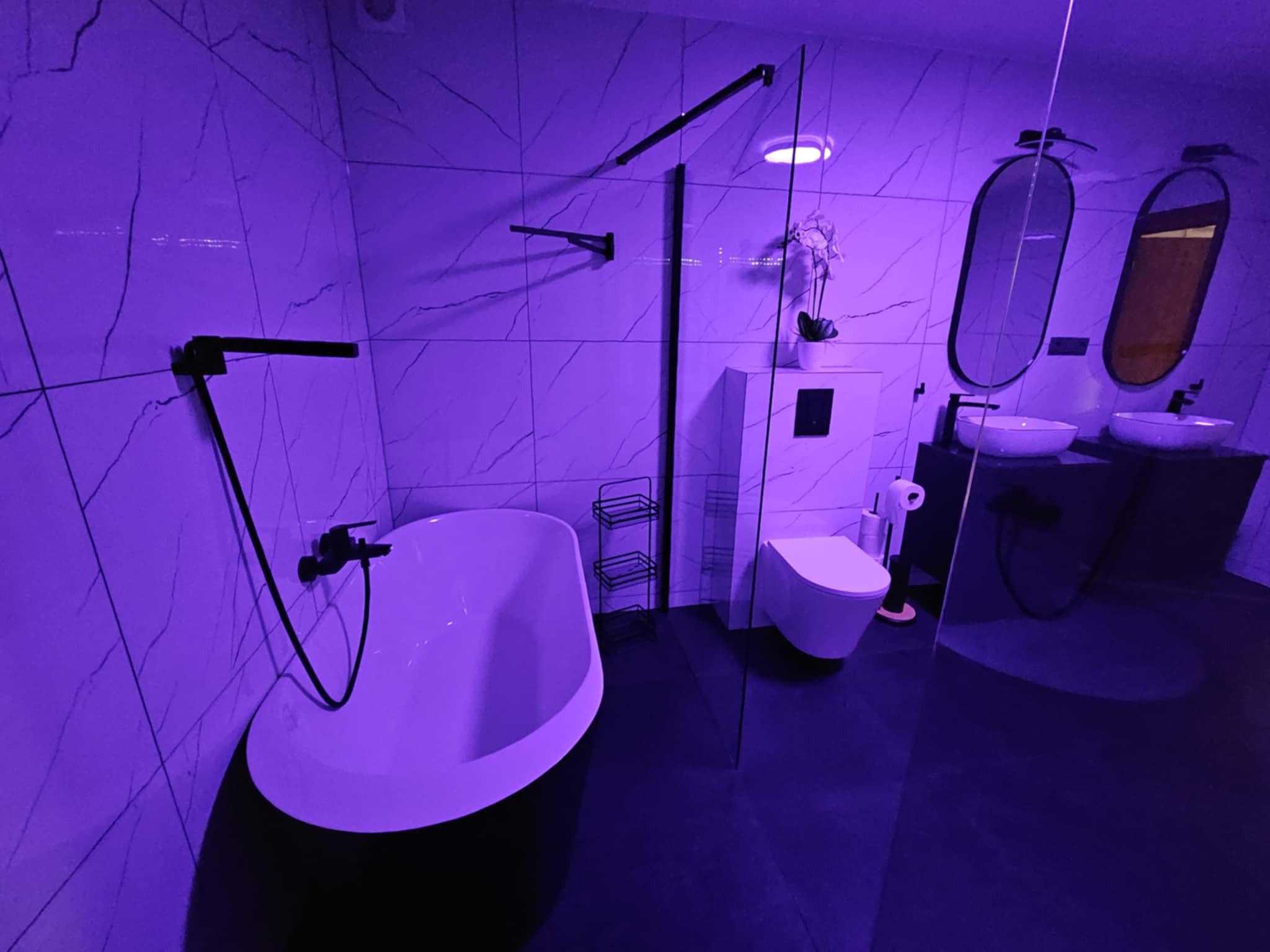 Nowoczesna łazienka z wolnostojącą wanną, umywalkami nablatowymi i marmurowymi płytkami ściennymi, oświetlona fioletowym światłem.