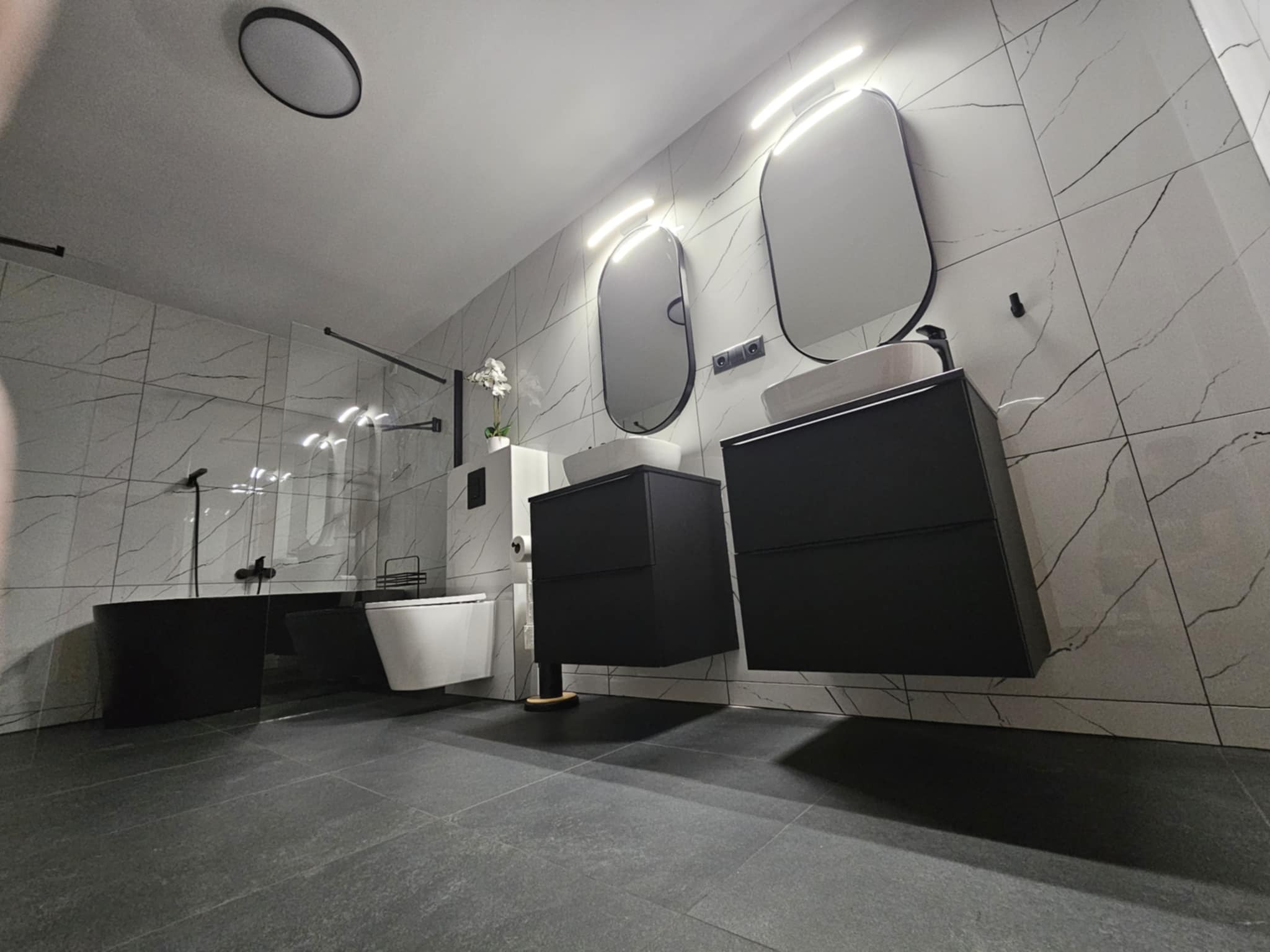 Nowoczesna łazienka z czarną wanną wolnostojącą, wiszącymi szafkami z umywalkami i białymi płytkami imitującymi marmur.