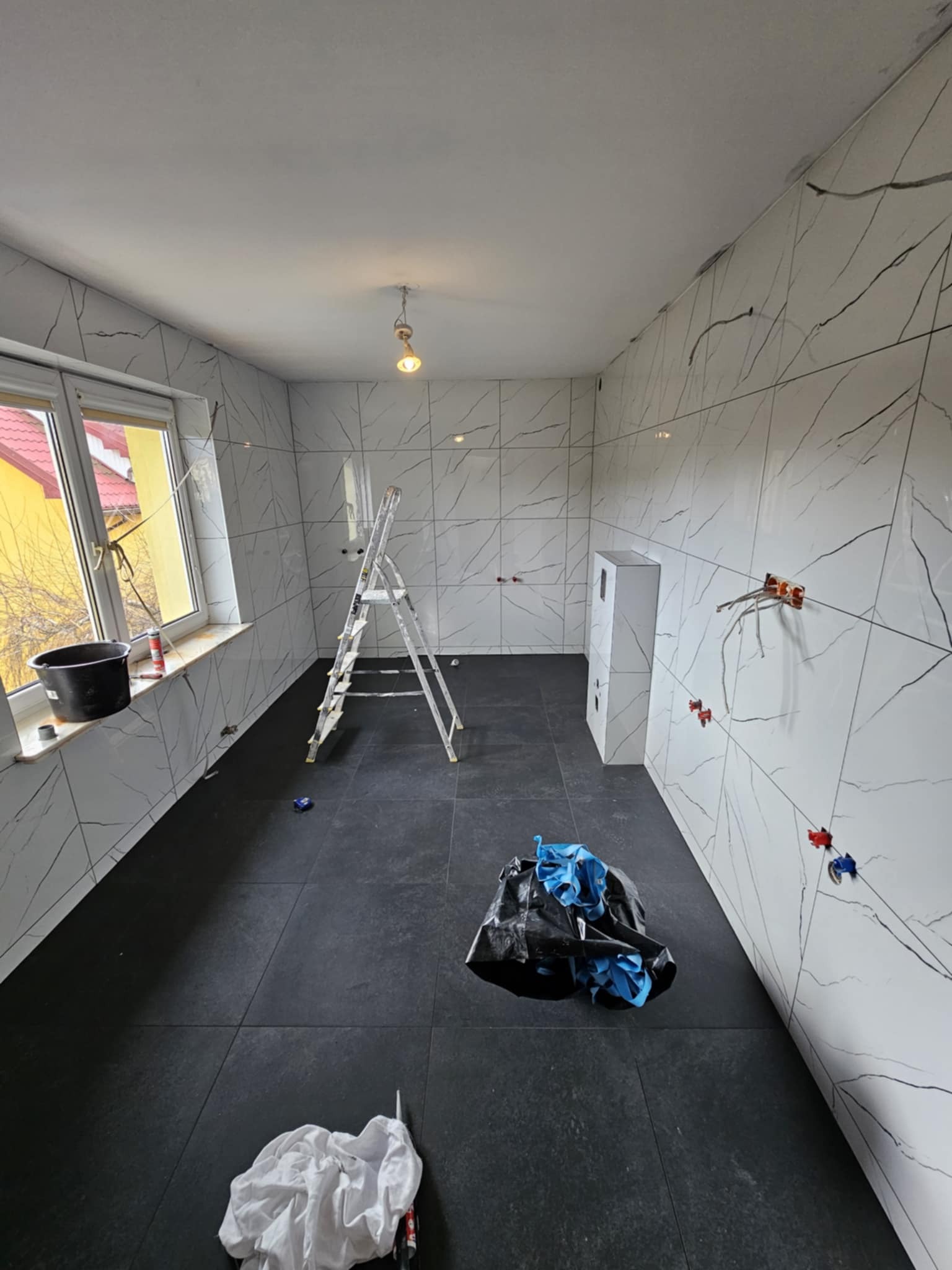 Wnętrze łazienki w trakcie remontu: białe płytki imitujące marmur z czarnymi żyłami, czarna podłoga, drabina aluminiowa, wystające kable elektryczne, śmieci w worku, widok z góry na pomieszczenie.