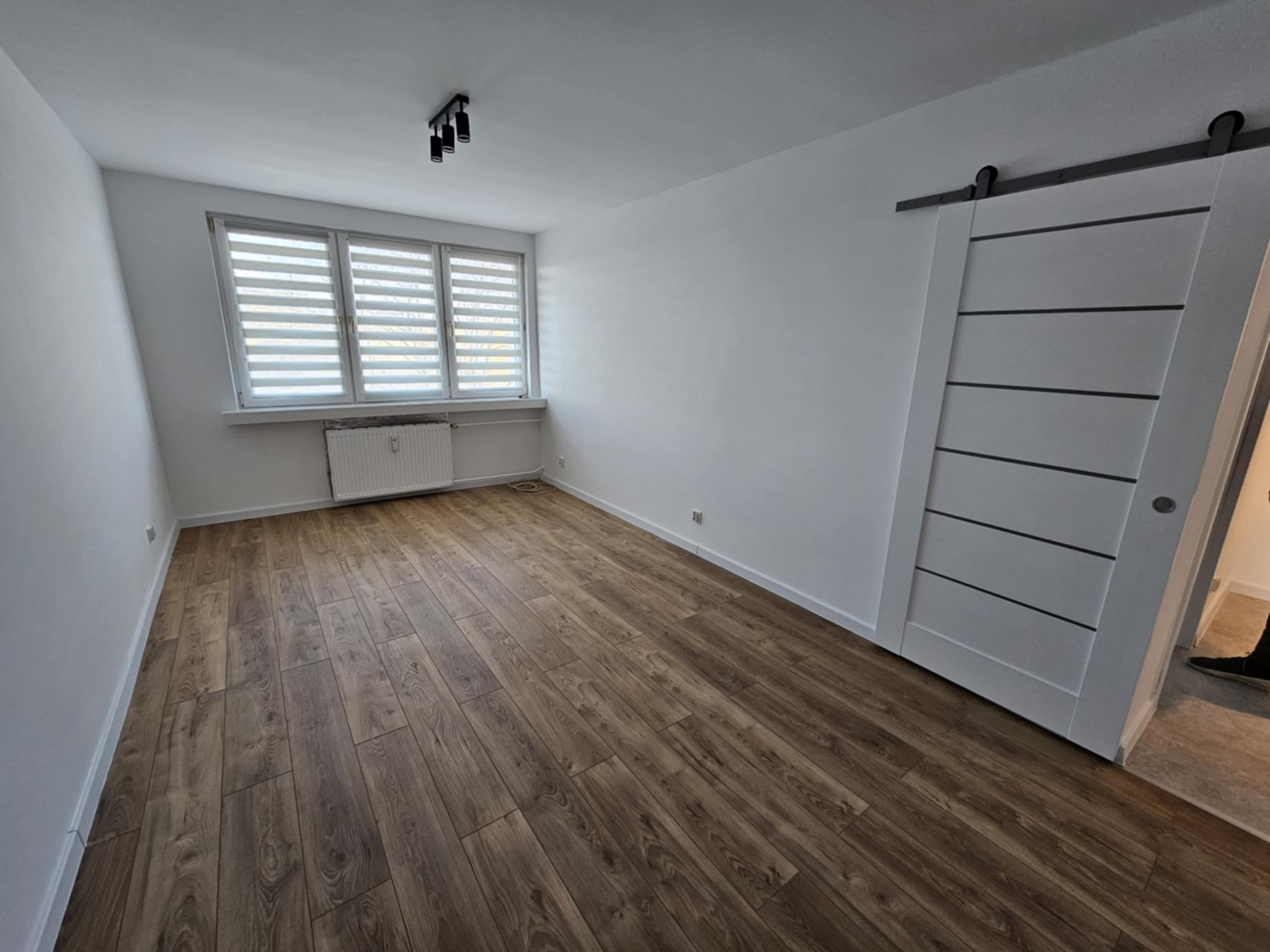 Pokój po remoncie z brązowymi panelami podłogowymi, białymi ścianami, grzejnikiem pod oknem z żaluzjami poziomymi i przesuwnymi drzwiami w stylu stodoły z czarnymi akcentami.