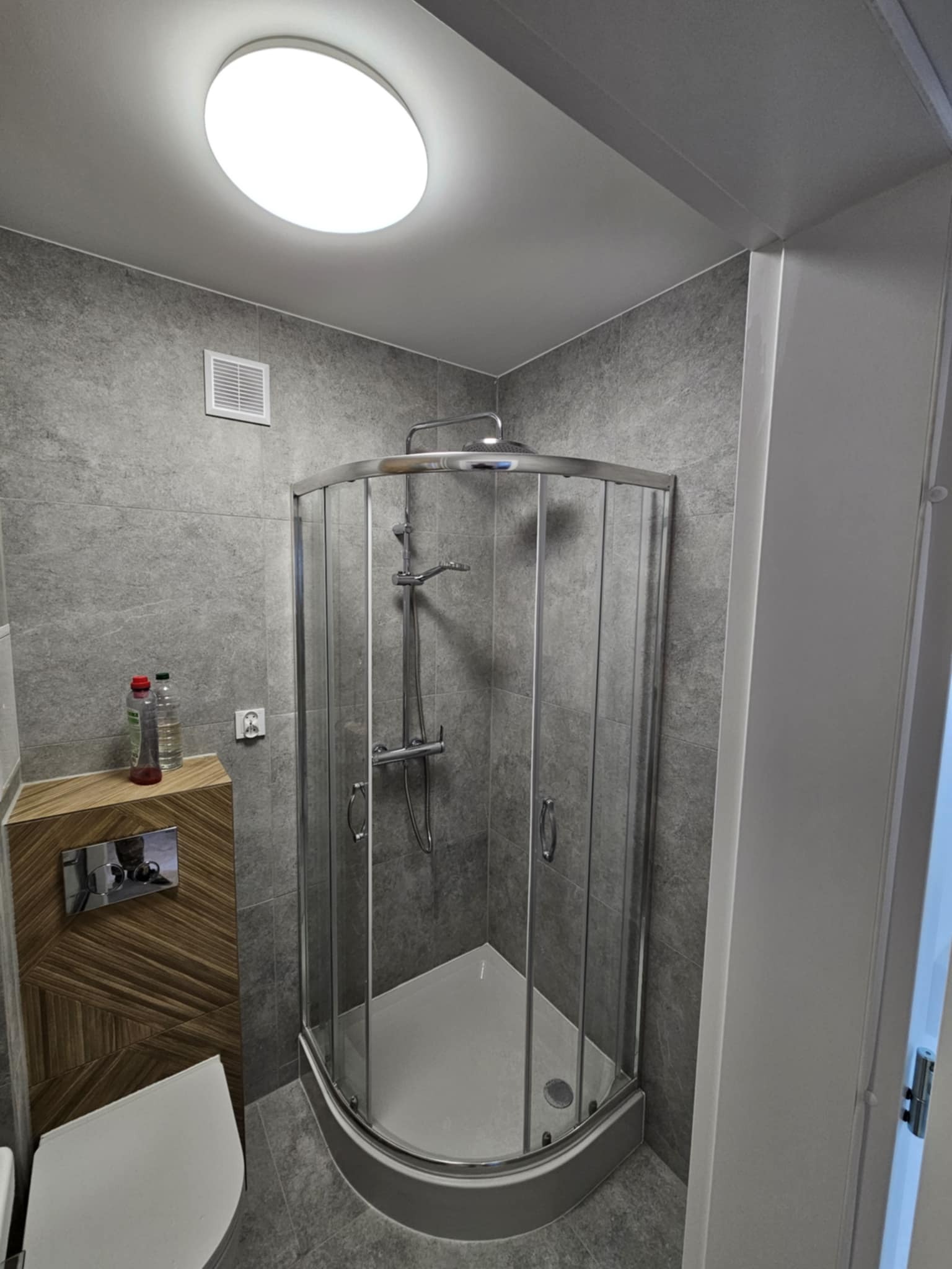 Wykończona łazienka w Koninie z kabiną prysznicową narożną, szarymi płytkami imitującymi kamień i zabudową toalety z drewnopodobnym wzorem w jodełkę.