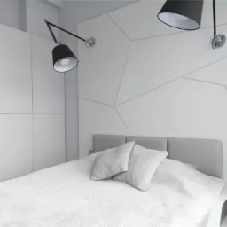 Minimalistyczna sypialnia w bieli i szarości z geometryczną ścianą za łóżkiem, oświetlona regulowanymi lampami ściennymi i wyposażona w szafy wnękowe z gładkimi frontami.