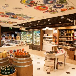 Wnętrze sklepu z alkoholem i delikatesami, z elementami dekoracyjnymi inspirowanymi folklorem, widoczne beczki z napojami i regały wypełnione butelkami.