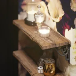 Dwu-półkowa, rustykalna półka z dekoracjami: świecami w szklanych pojemnikach, złotą szkatułką i biało-złotym pudełeczkiem, na tle ściany w kwiatowe tapety w odcieniach fioletu i bieli.