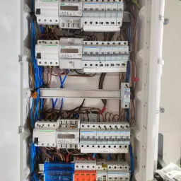 Rozdzielnica elektryczna z widocznymi zabezpieczeniami, licznikami energii i okablowaniem w kolorach niebieskim, brązowym i ż&oacute;łto-zielonym, zamontowana w białej obudowie, z widocznymi elementami...