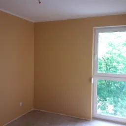 Pomalowane na żółto ściany pokoju z białym oknem balkonowym, widok na zieleń za oknem, wykończona listwa przypodłogowa.