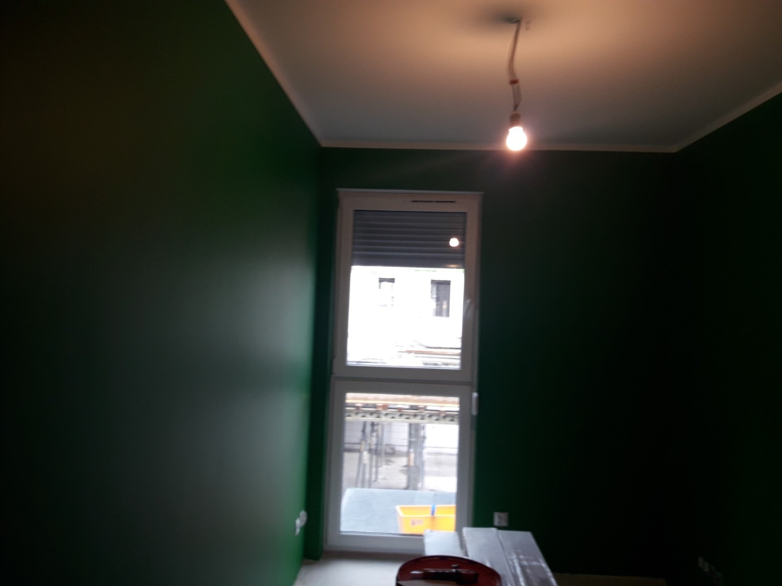 Świeżo pomalowane na ciemnozielono ściany w pokoju z oknem i widoczną żarówką na suficie, częściowo zakryte materiały malarskie na podłodze.
