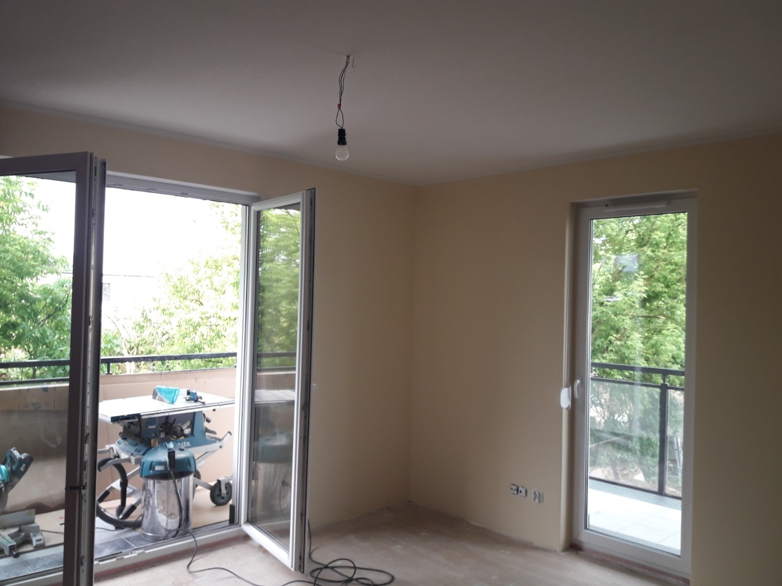 Wnętrze pokoju w trakcie remontu z otwartym balkonem, widoczna piła stołowa i odkurzacz przemysłowy, surowe ściany w kolorze beżowym, wisząca żarówka bez klosza.