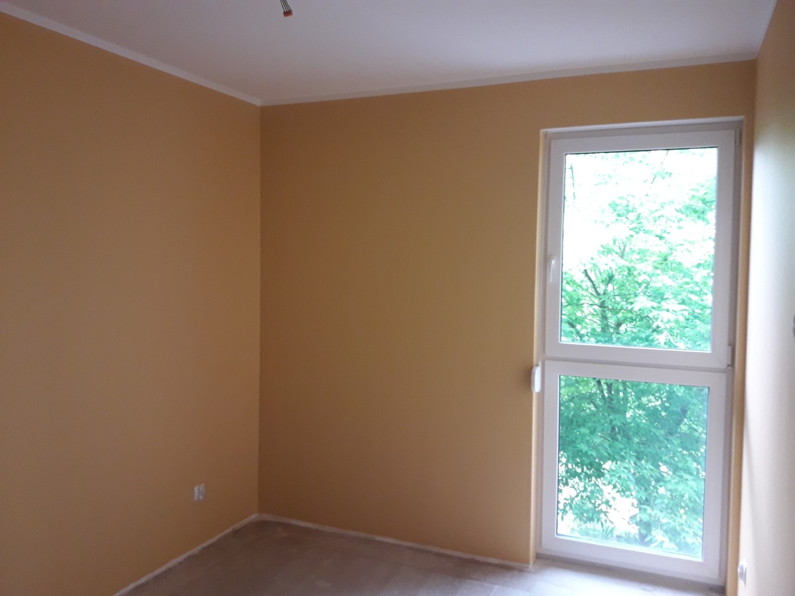 Pomalowane na żółto ściany pokoju z białym oknem balkonowym, widok na zieleń za oknem, wykończona listwa przypodłogowa.