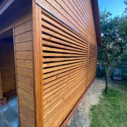 Zewnętrzny widok nowej, drewnianej altany z poziomymi, ażurowymi panelami, osadzonej na ceglanej podmurówce, na tle zielonego trawnika i drzew.