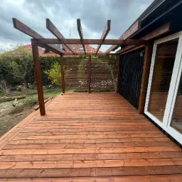 Nowoczesny taras z drewna o ciepłym, brązowym odcieniu, z drewnianą pergolą i czarną ścianą budynku, widok na ogród z magnolią w tle.