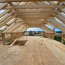 Patrick Design Apartments - Wnętrze domu w trakcie budowy, widok na drewnianą konstrukcję dachu i podłogi. W tle zabudowa jednorodzinna i błękitne niebo.