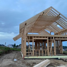 Patrick Design Apartments - Surowa konstrukcja domu szkieletowego z drewna na tle pochmurnego nieba. Widoczne elementy konstrukcyjne i teren budowy. W tle zabudowa jednorodzinna.