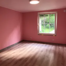 Pusty pokój z różowymi ścianami, panelami podłogowymi w odcieniach brązu i jasnym oknem z widokiem na zieleń.
