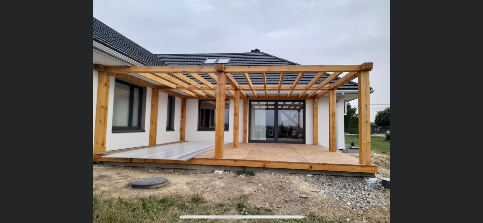Drewniana pergola zadaszająca taras przy nowoczesnym domu z ciemną dachówką, widoczne częściowo zamontowane panele z tworzywa sztucznego, otoczenie w trakcie prac wykończeniowych.