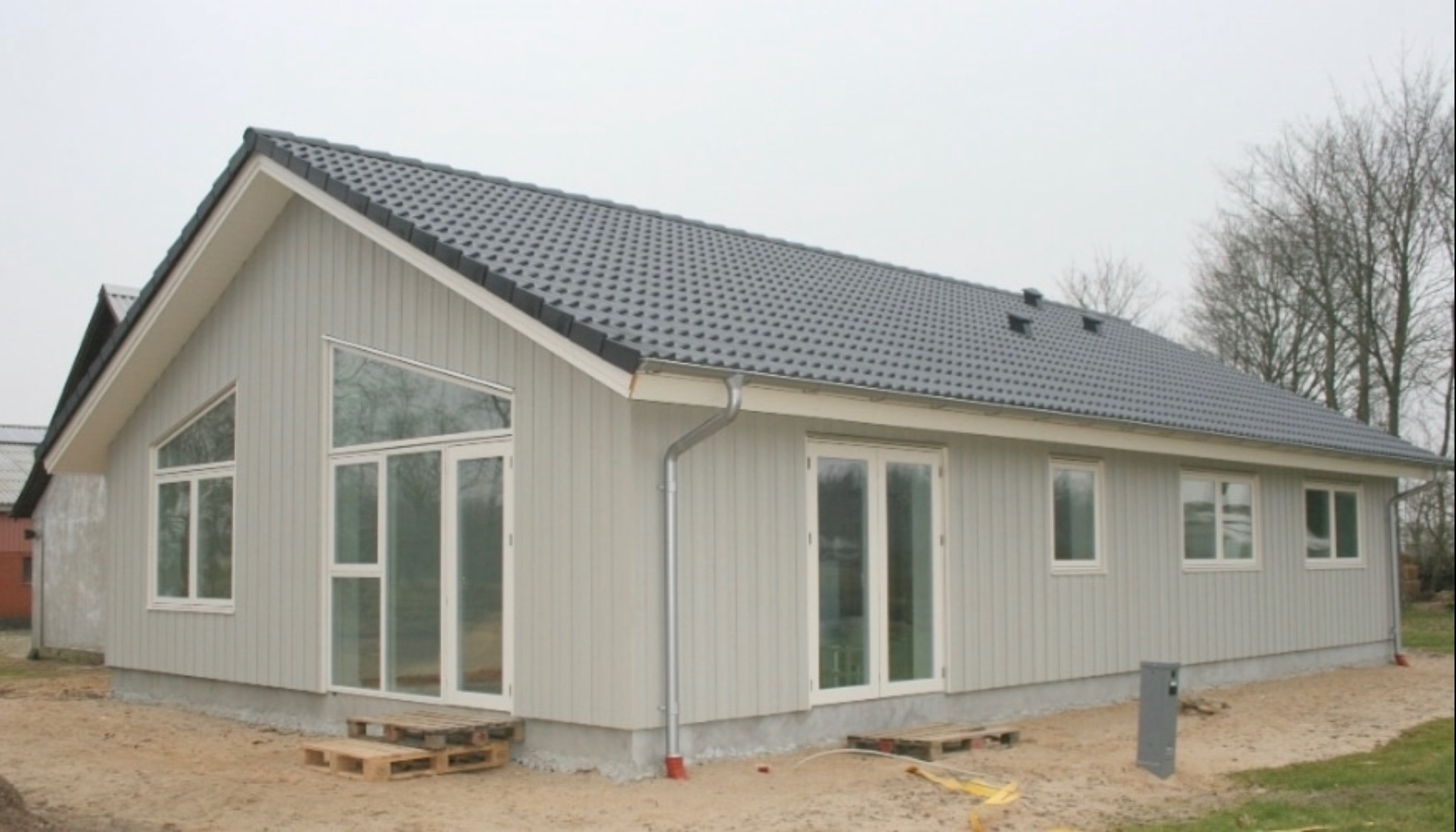 Gotowy, parterowy dom z szarej deski z ciemnoszarą dachówką, widoczne palety przed wejściem i rynna odprowadzająca wodę.