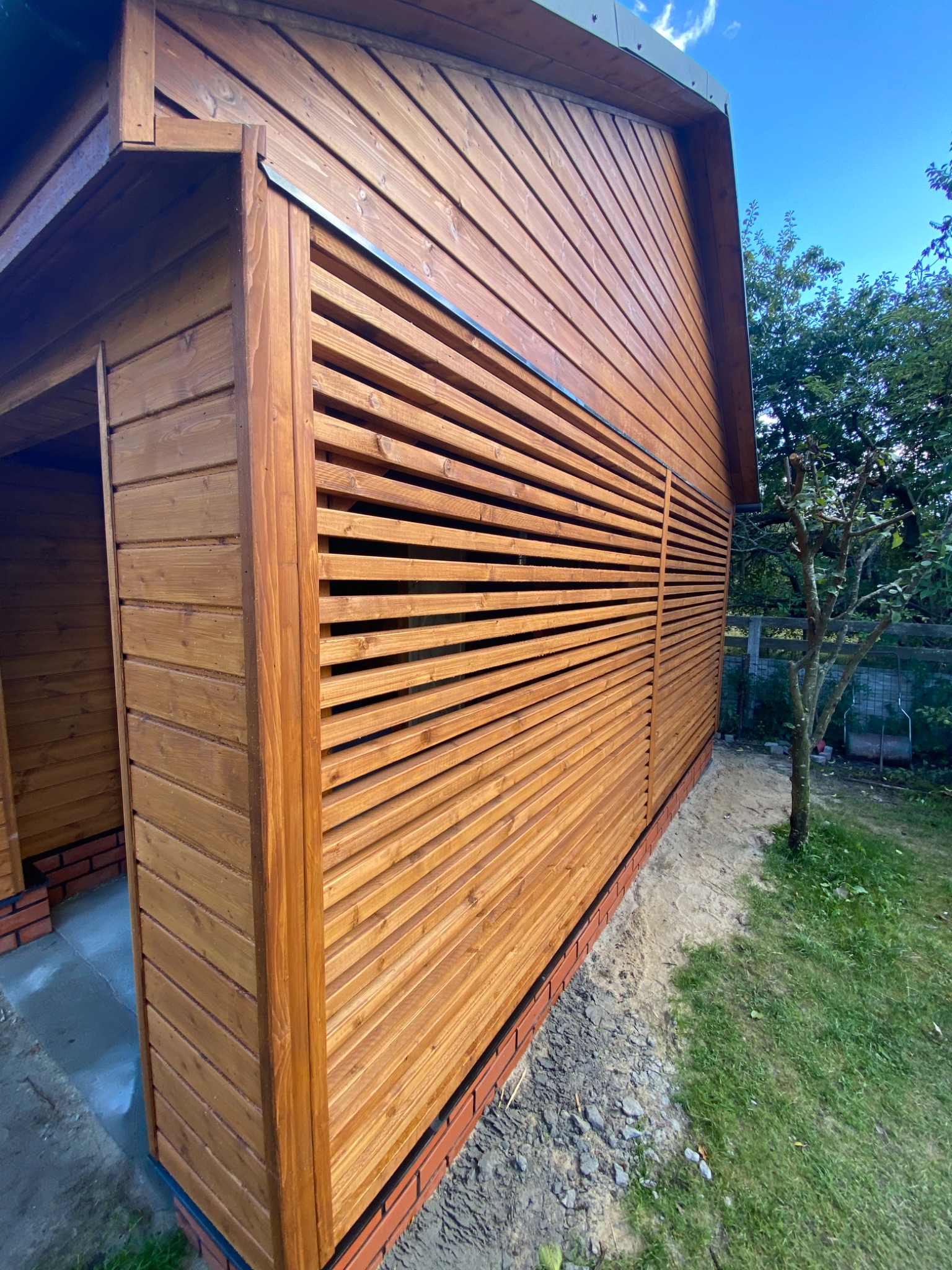 Zewnętrzny widok nowej, drewnianej altany z poziomymi, ażurowymi panelami, osadzonej na ceglanej podmurówce, na tle zielonego trawnika i drzew.