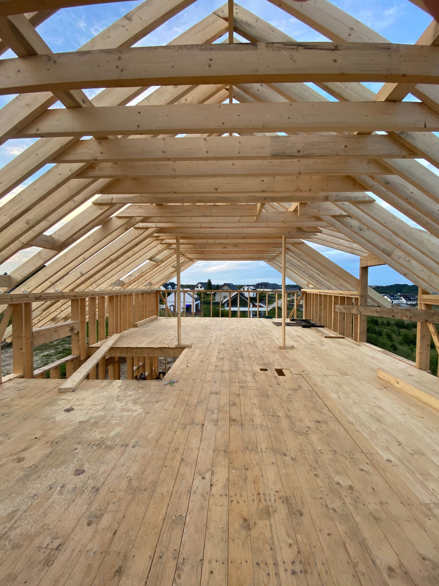 Wnętrze domu w trakcie budowy, widok na drewnianą konstrukcję dachu i podłogi. W tle zabudowa jednorodzinna i błękitne niebo.