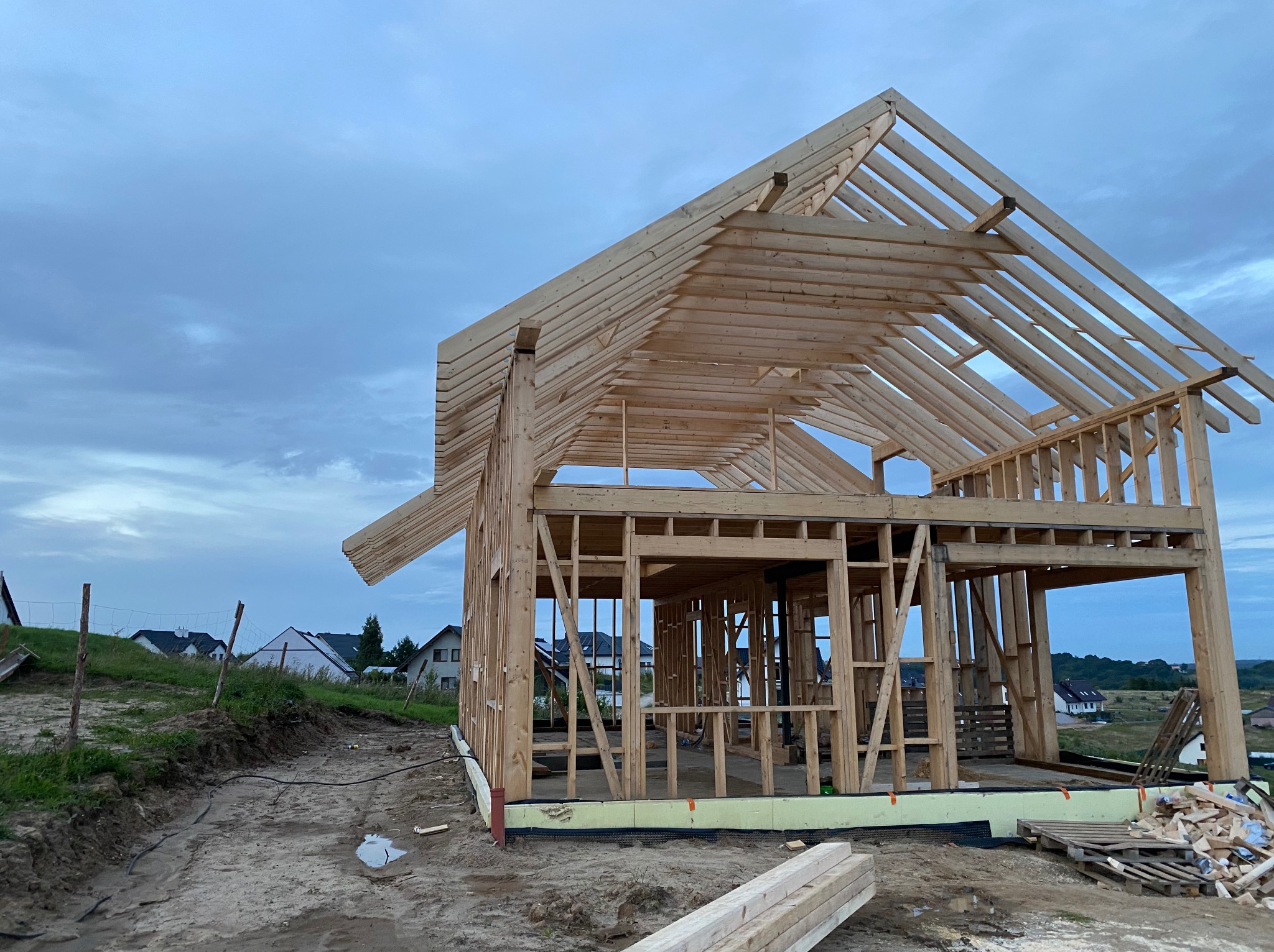 Surowa konstrukcja domu szkieletowego z drewna na tle pochmurnego nieba. Widoczne elementy konstrukcyjne i teren budowy. W tle zabudowa jednorodzinna.