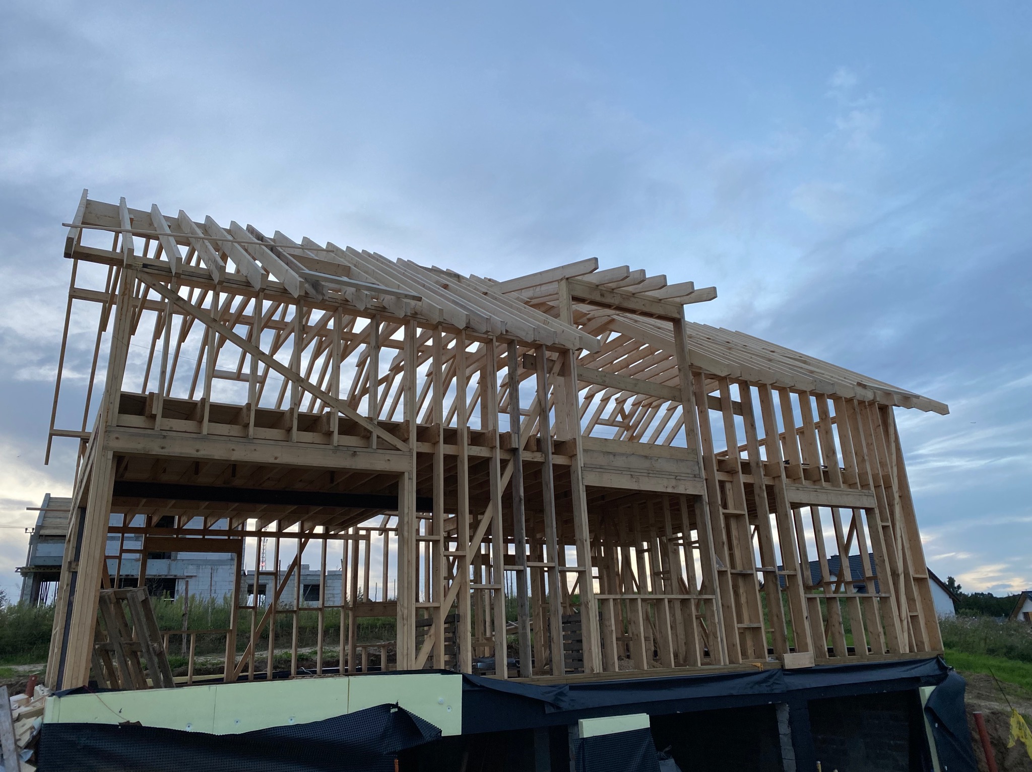 Surowa konstrukcja domu szkieletowego z widocznym drewnianym szkieletem ścian i dachu na tle pochmurnego nieba. Widoczna izolacja fundamentów i sąsiedni budynek w tle.