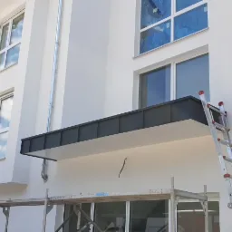 Nowoczesny, minimalistyczny daszek nad wejściem do budynku, z czarnym obramowaniem i białym podsufitką, wsparty na metalowych rurach, widoczny z rusztowania i drabiny.