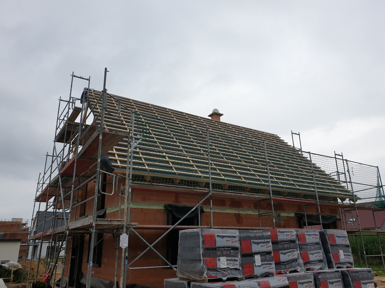 Dom w trakcie budowy z widoczną konstrukcją więźby dachowej, rusztowaniami i paletami z cegłą klinkierową Hagemester na pierwszym planie.