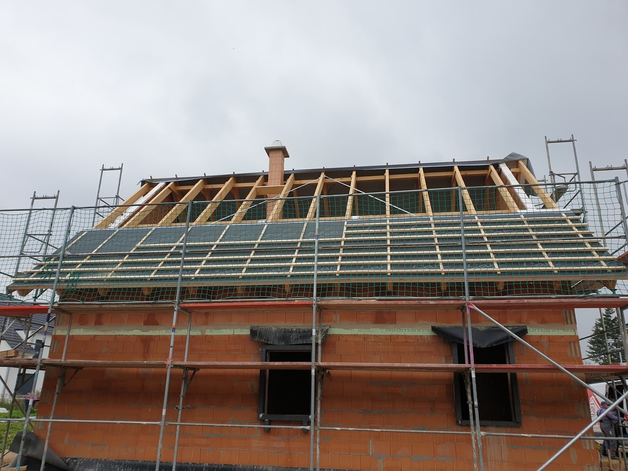 Wymiana dachu w trakcie: widok na konstrukcję drewnianą, membranę dachową i rusztowanie przy ceglanym budynku z kominem, pochmurne niebo.