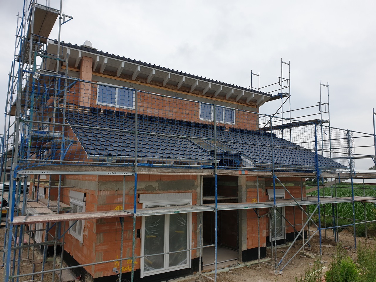 Dom w trakcie budowy, widoczne rusztowania, cegły i częściowo położony dach z ciemnej dachówki. Widoczna siatka zabezpieczająca na rusztowaniach.