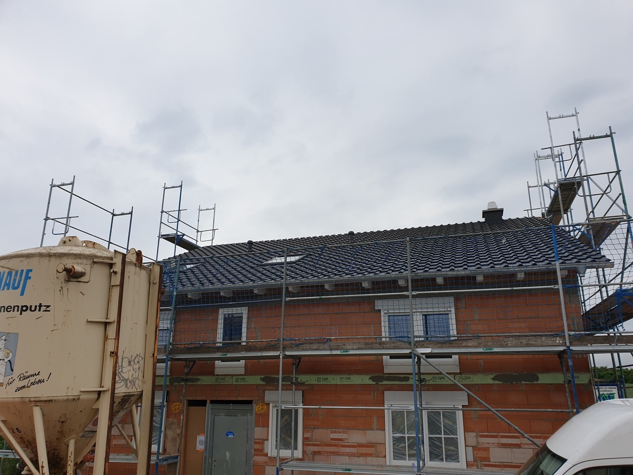 Dom w trakcie budowy z częściowo położonym, ciemnym dachem, otoczony rusztowaniami i zabezpieczony siatką, z widocznym zbiornikiem na zaprawę Knauf.