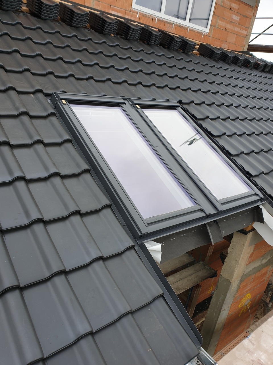 Nowy dach z czarnej dachówki ceramicznej z dwoma oknami dachowymi, widoczne elementy konstrukcji i fragment muru z cegły