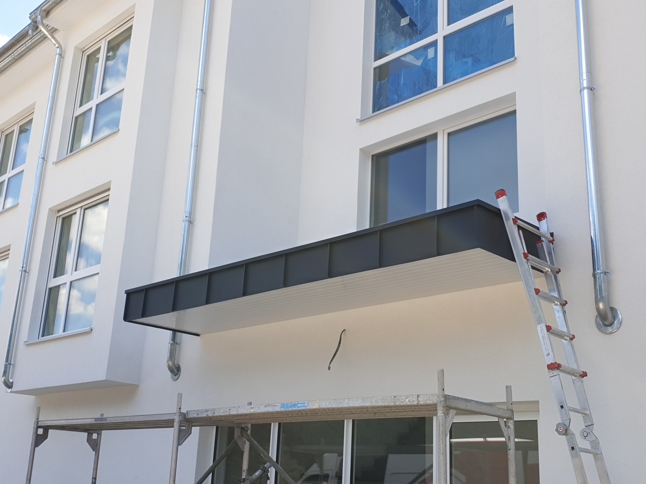 Nowoczesny, minimalistyczny daszek nad wejściem do budynku, z czarnym obramowaniem i białym podsufitką, wsparty na metalowych rurach, widoczny z rusztowania i drabiny.