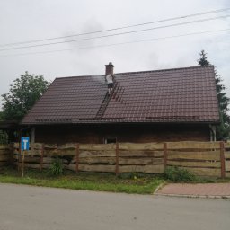 Firma Usługowa Konstanty Romankiewicz - Mężczyzna na drabinie czyści dach brązowego domu z kominem, widoczne strumienie wody i drewniane ogrodzenie.