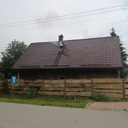 Mężczyzna na drabinie czyści dach brązowego domu z kominem, widoczne strumienie wody i drewniane ogrodzenie.