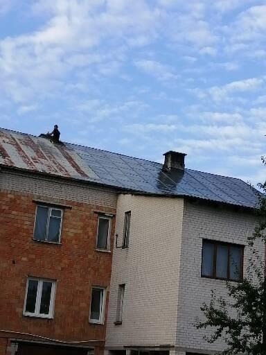 Dwupiętrowy budynek z częściowo odnowionym dachem; po lewej stronie stara, rdzawa blacha, po prawej nowa, ciemna dachówka; na dachu, przy kominie, sylwetka pracownika w ciemnym ubraniu.