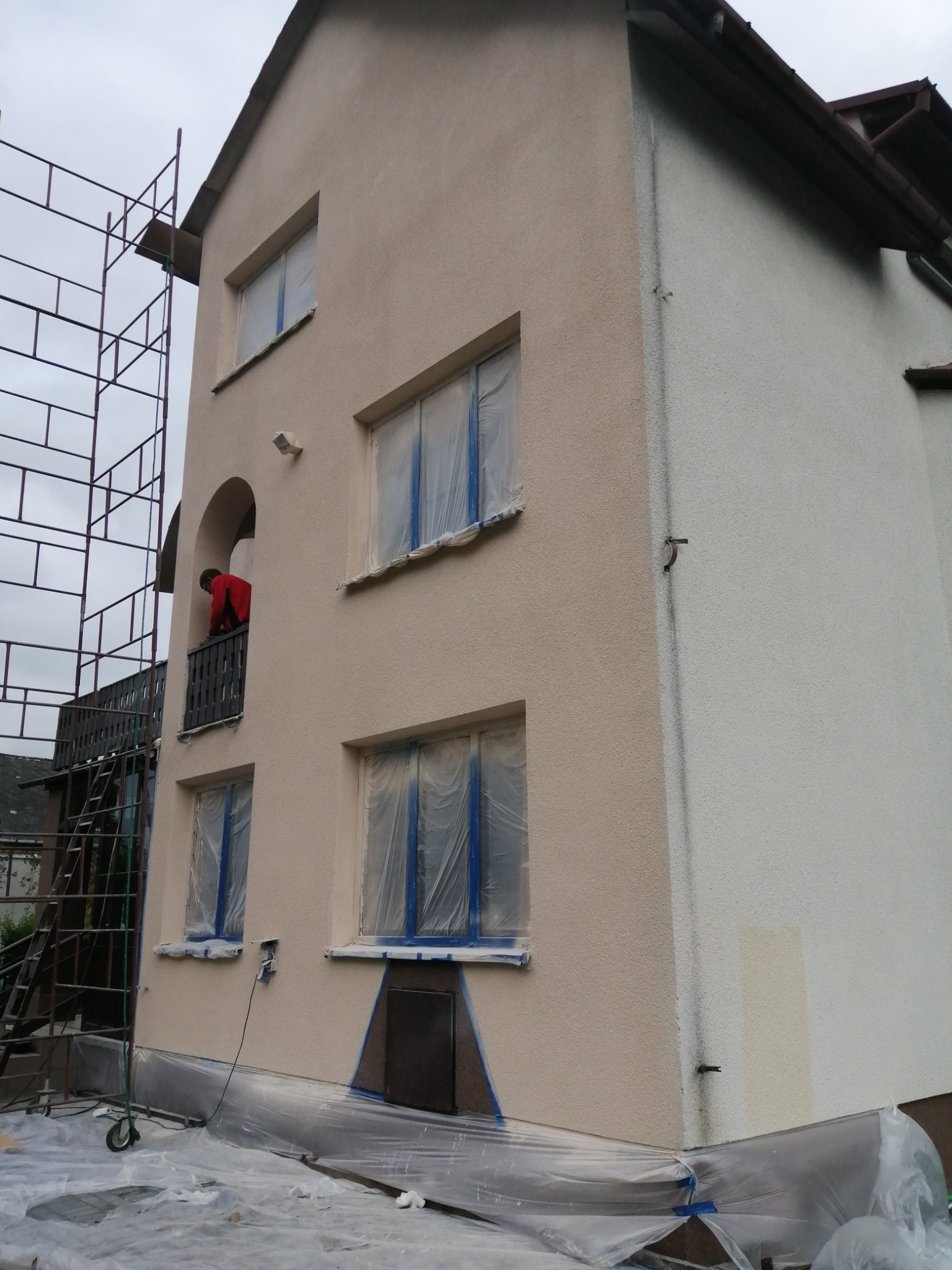 Dwukondygnacyjny dom z beżową i białą elewacją w trakcie prac, okna zabezpieczone folią, widoczne rusztowanie i pracownik w czerwonej bluzie na balkonie.