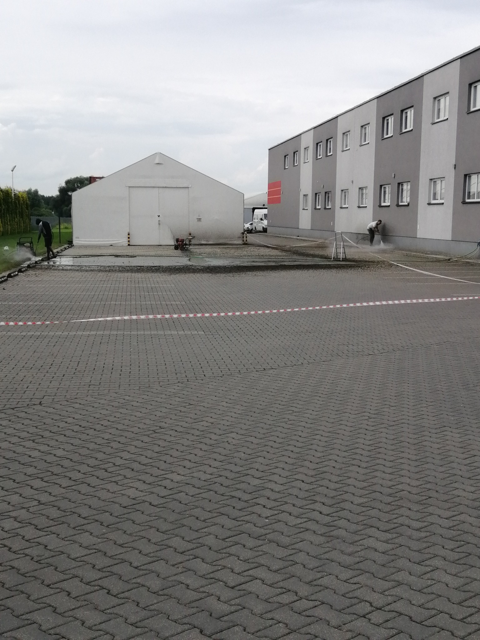 Pracownicy czyszczący elewację budynku i kostkę brukową za pomocą myjek ciśnieniowych, widoczny biały magazyn w tle, pochmurne niebo.