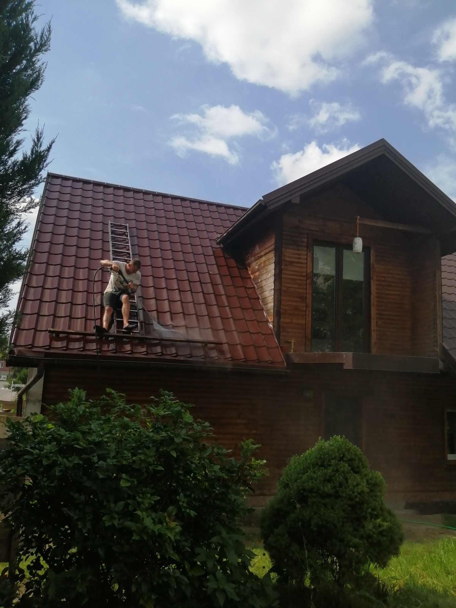 Pracownik na drabinie czyści dach z brązowej dachówki za pomocą myjki ciśnieniowej; widoczny drewniany dom z oknami i zielona roślinność.