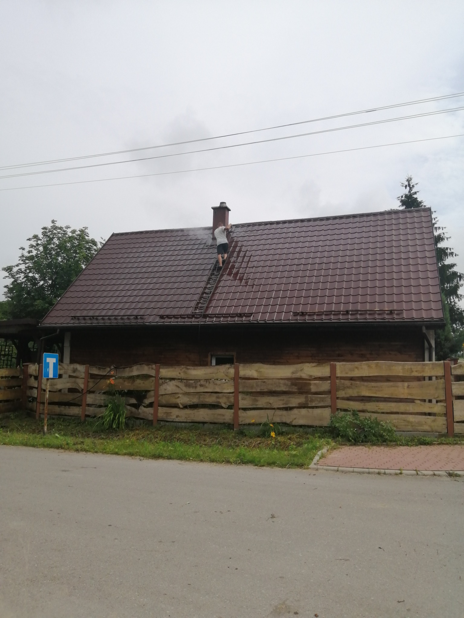 Mężczyzna na drabinie czyści dach brązowego domu z kominem, widoczne strumienie wody i drewniane ogrodzenie.