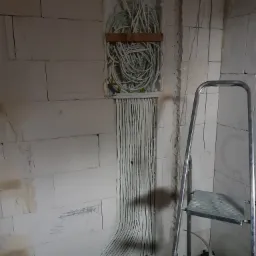 Rozprowadzenie kabli elektrycznych w ścianie z betonu komórkowego, widoczne kable w peszlach i puszka instalacyjna z wiązką przewodów, drabina aluminiowa w tle.