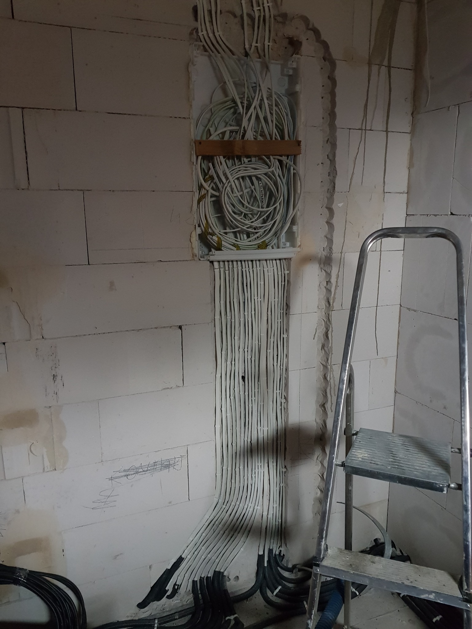 Rozprowadzenie kabli elektrycznych w ścianie z betonu komórkowego, widoczne kable w peszlach i puszka instalacyjna z wiązką przewodów, drabina aluminiowa w tle.
