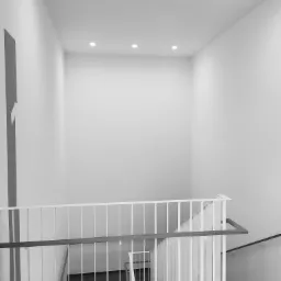 Biało-szare wnętrze klatki schodowej z minimalistyczną balustradą i punktowym oświetleniem sufitowym.