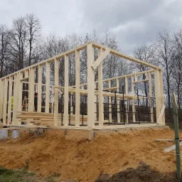 Wznoszenie drewnianej konstrukcji budynku na fundamencie w trakcie budowy, widoczne elementy szkieletu, pracownicy w odblaskowych kamizelkach i drzewa w tle.