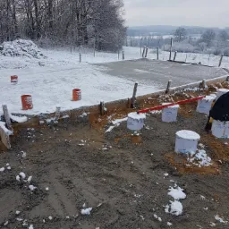 Zimowy krajobraz budowy fundamentów: widoczne szalunki, przygotowane podpory zalane betonem, częściowo odśnieżony teren i zaśnieżone drzewa w tle.