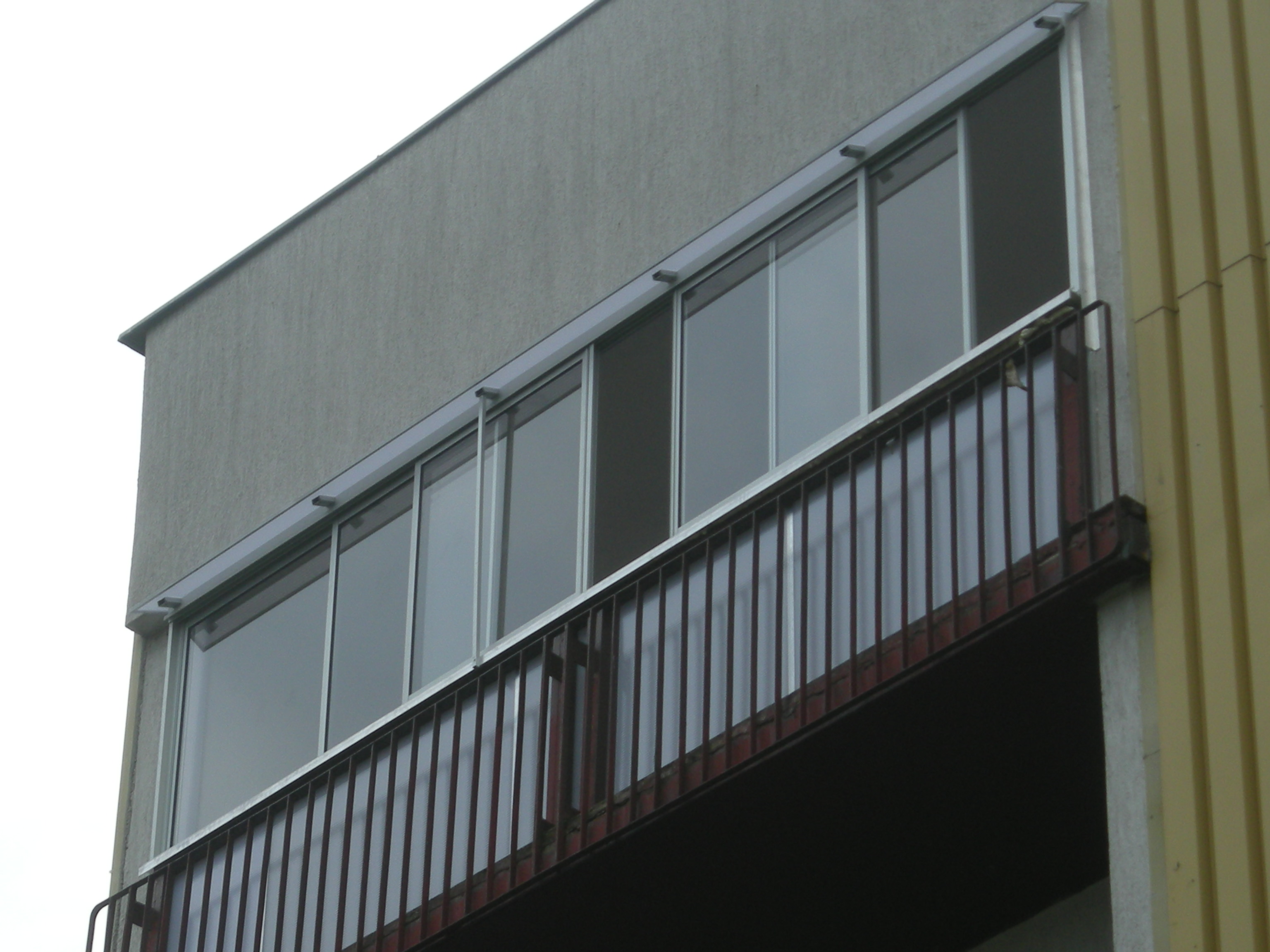 Zabudowany balkon z aluminiową stolarką okienną, widok z dołu na fragment budynku z szarą elewacją i metalową barierką balkonu.