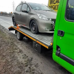 Szary samochód osobowy zabezpieczony pomarańczowymi pasami na platformie lawety, widok z boku, z zielonym bokiem pojazdu lawety po prawej stronie, w tle żółty billboard.