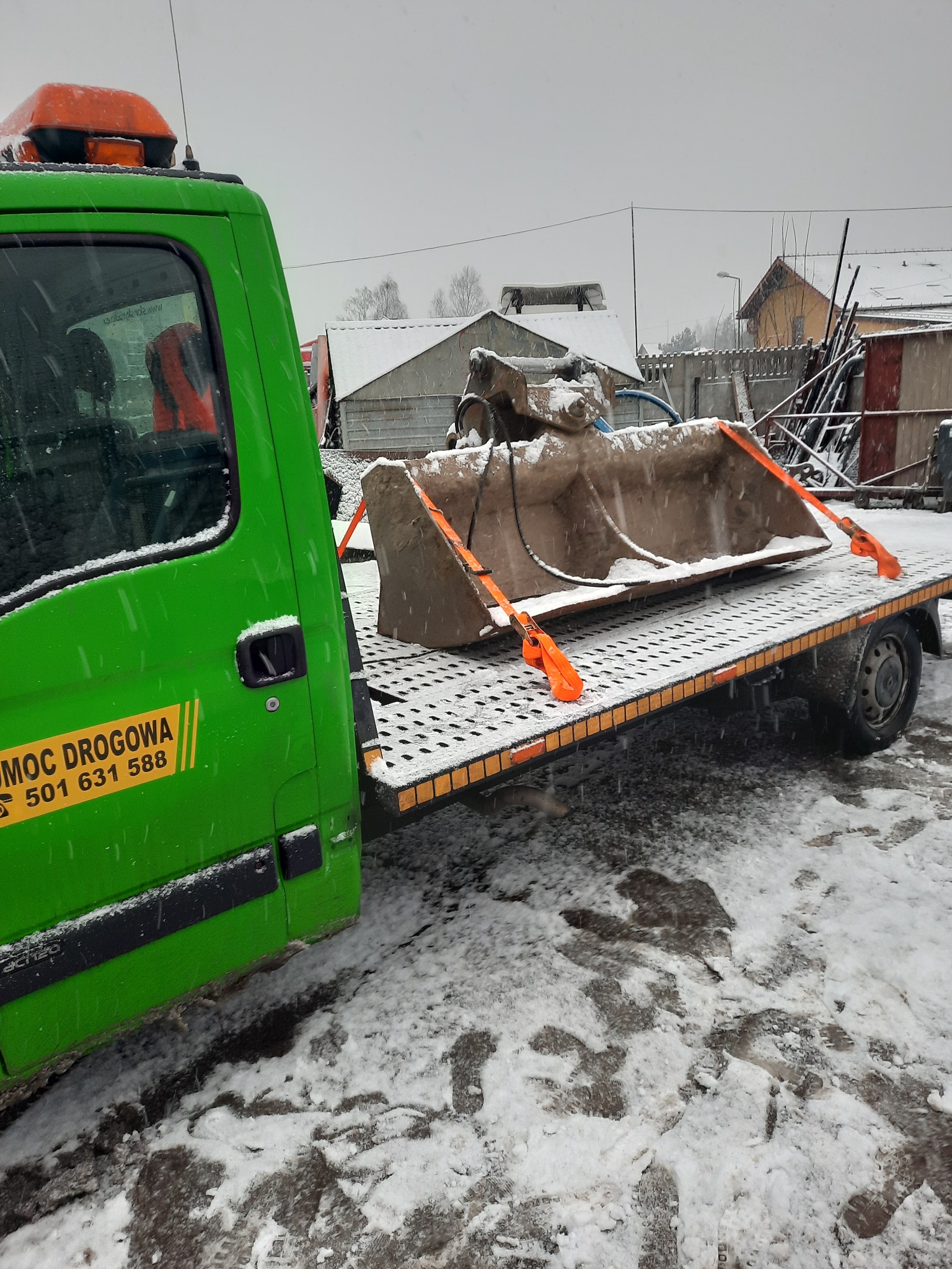 Zielony samochód pomocy drogowej z pomarańczowym kogutem przewozi łyżkę koparki zabezpieczoną pomarańczowymi pasami transportowymi podczas śnieżnej pogody.