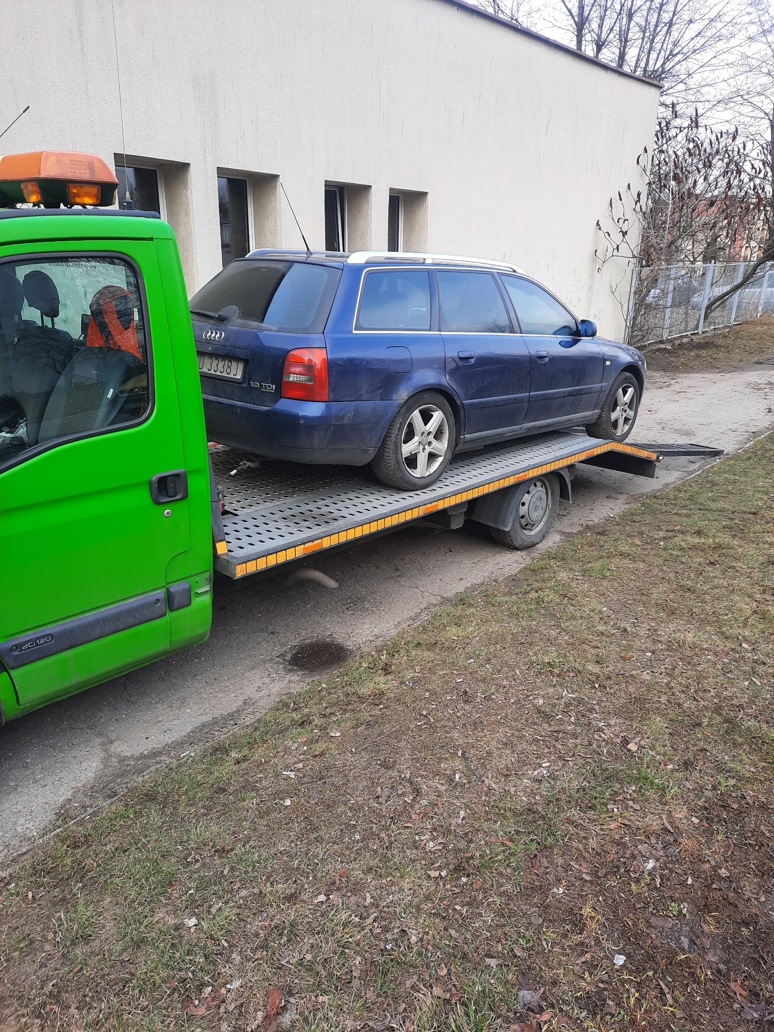 Zielony holownik z pomarańczowym kogutem przewozi granatowe kombi Audi na platformie. Widoczne budynki w tle.