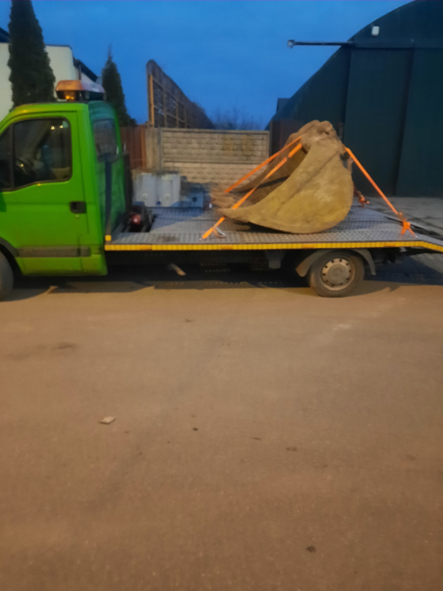 Zielony samochód laweta z pomarańczowym kogutem przewozi łyżkę koparki zabezpieczoną pomarańczowymi pasami.